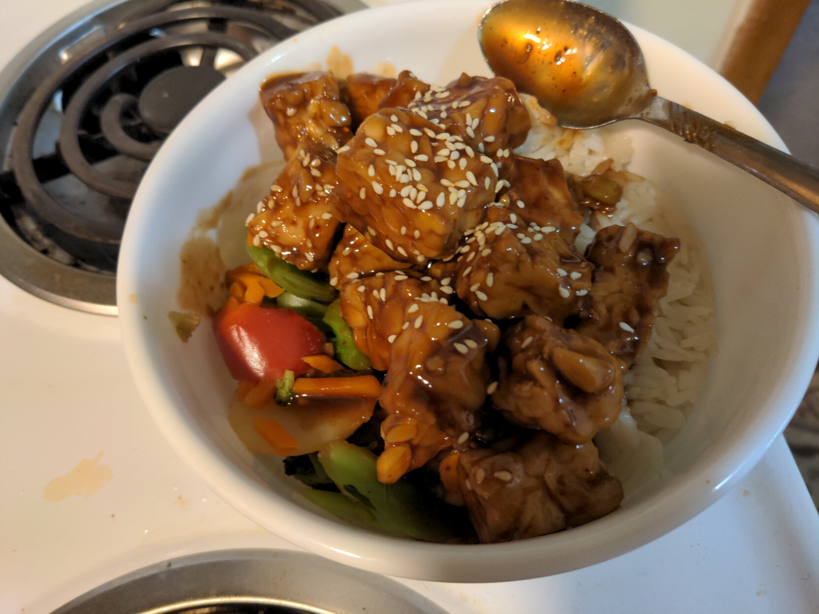 A bowl of stir fry vegetables and jasmine rice, topped with saucy tempeh and garnished with white sesame seeds.